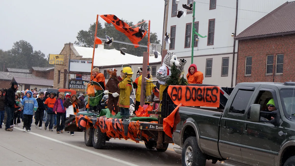 Small Town Homecoming Parade Float on a Flatbed Trailer Homecoming parade float on a flatbed trailer pulled by a pickup down a small-town main street, with an orange banner, costumed riders and spectators