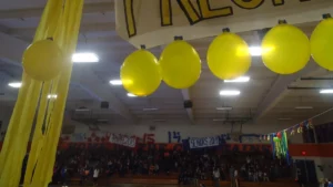 Yellow balloons and a freshman class banner hanging over packed gym bleachers with senior class signs on the far wall before pep rally games