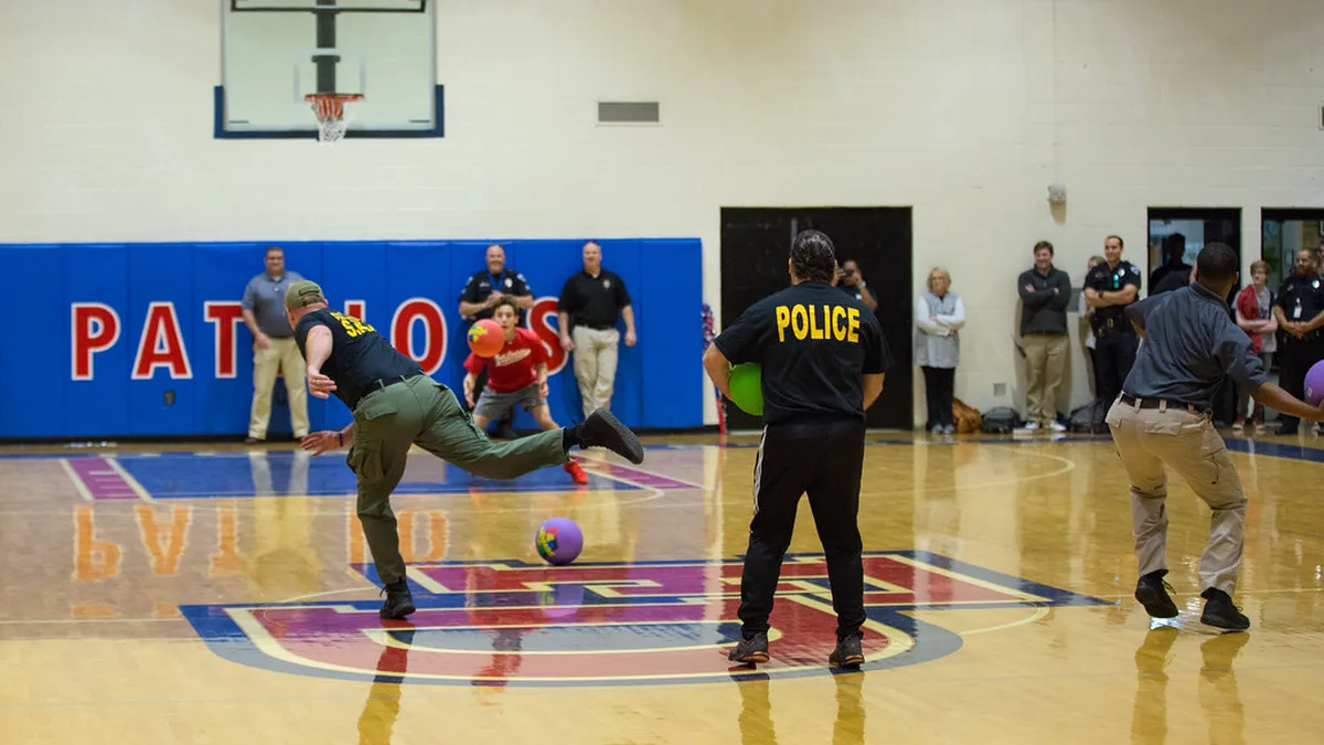Staff vs Students Style Dodgeball Game in a High School Gym Adults in police shirts rolling and throwing soft balls across a high school gym floor while students and staff watch from the baseline