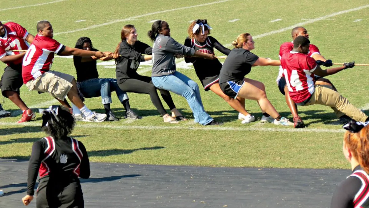 Tug of War Between Students and Staff at an Outdoor Pep Rally A mixed team of students in jerseys and adults pulling a rope in a tug of war on a football field while cheerleaders watch at a pep rally