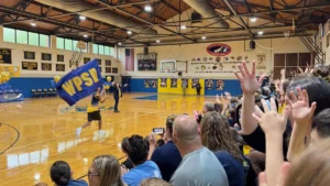 Crowd in a school gymnasium cheering as a student runs the school flag past balloons in school colors at a homecoming pep rally