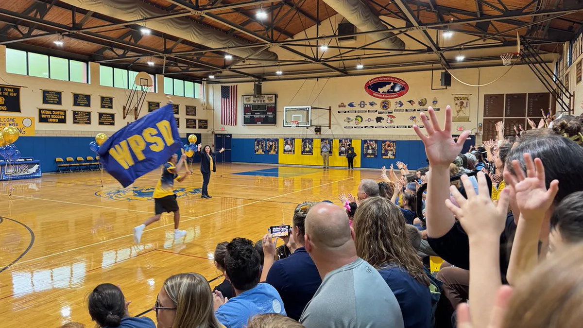 Homecoming Pep Rally in a School Gym During Spirit Week Crowd in a school gymnasium cheering as a student runs the school flag past balloons in school colors at a homecoming pep rally